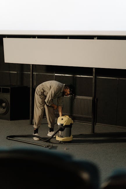 A person in a light-colored work uniform is operating a portable industrial vacuum cleaner on a carpeted floor of an indoor space, likely a conference or event hall. The individual is bent over, focus