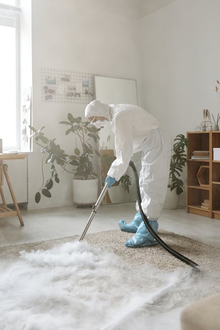 A person in a light-colored work uniform is operating a portable industrial vacuum cleaner on a carpeted floor of an indoor space, likely a conference or event hall. The individual is bent over, focus